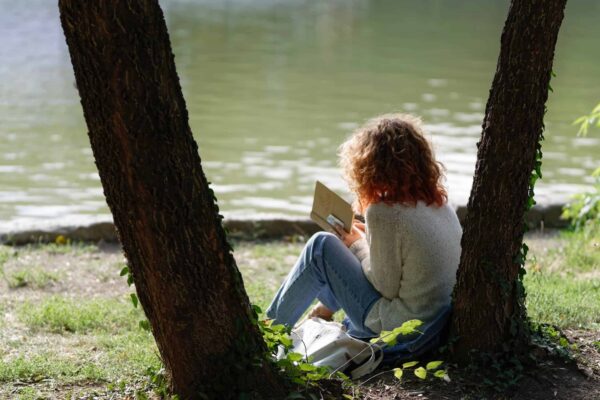 girl with red hair reading a book by a body of water